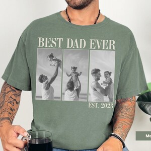 Moss green Comfort Colors t-shirt worn by a man holding a mug, with bold “BEST DAD EVER” text and a black-and-white three-photo collage of a dad with his child, labeled “EST. 2023.” Features sweet candid moments of fatherhood. Personalized shirt perfect for first-time dads, Father’s Day, or baby’s first birthday. A heartfelt and stylish custom gift celebrating dad life.