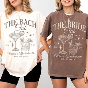May include: Two women wearing t-shirts with a bachelorette party design. The shirts are in ivory and espresso colors. The design features the text "The Bach Club" and "The Bride Club" with a cocktail glass and a glass of ice. The text also includes "Est. 2025" and "Camila's Bachelorette, Palm Springs, CA".