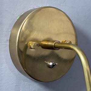 May include: A brass wall lamp featuring a circular base and a curved arm. The base includes a small switch and a knurled adjustment knob. The lamp is mounted on a light grey wall.