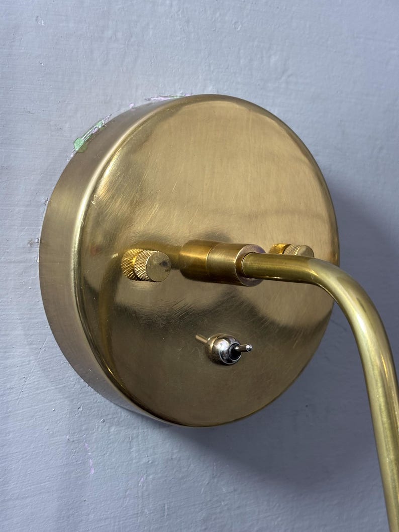 May include: A brass wall lamp featuring a circular base and a curved arm. The base includes a small switch and a knurled adjustment knob. The lamp is mounted on a light grey wall.