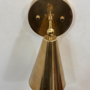 May include: A brass wall sconce featuring a conical shade and a circular backplate. The sconce has a polished finish and a simple, elegant design. The light fixture is mounted on a white wall.