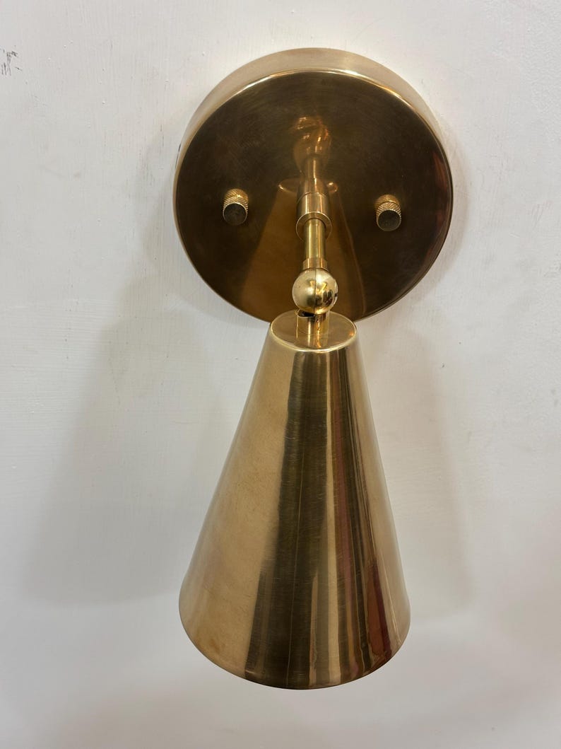 May include: A brass wall sconce featuring a conical shade and a circular backplate. The sconce has a polished finish and a simple, elegant design. The light fixture is mounted on a white wall.