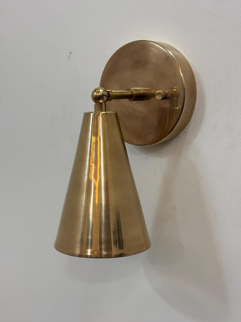 May include: A brass wall sconce featuring a conical lampshade and a circular backplate. The sconce has a warm, metallic finish and is mounted on a white wall. The design is simple and modern, suitable for interior lighting.