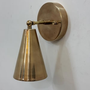 May include: A brass wall sconce featuring a conical shade and a circular backplate. The sconce has a warm, metallic finish and is mounted on a white wall. The design is minimalist and modern, suitable for interior lighting.