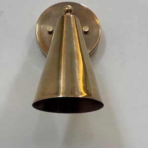 May include: A brass wall sconce featuring a conical shade and a circular backplate. The sconce has a polished finish and is mounted on a white wall. The design is minimalist and modern, suitable for interior lighting in the UK.