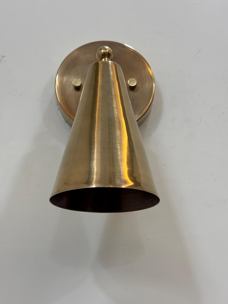 May include: A brass wall sconce featuring a conical shade and a circular backplate. The sconce has a polished finish and is mounted on a white wall. The design is minimalist and modern, suitable for interior lighting in the UK.