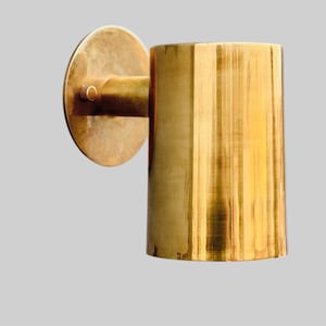 May include: A gold-colored wall sconce with a cylindrical shade and a circular mounting plate. The sconce is made of metal and has a brushed finish. The design is minimalist and modern, suitable for interior lighting.