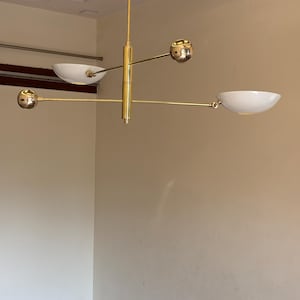 May include: A gold-toned modern chandelier with four white bowl-shaped light fixtures and four gold spheres. The chandelier has a central vertical rod with three horizontal arms extending out.