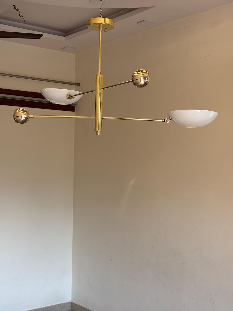 May include: A gold-toned modern chandelier with four white bowl-shaped light fixtures and four gold spheres. The chandelier has a central vertical rod with three horizontal arms extending out.
