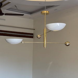 May include: A gold-toned modern chandelier with three white bowl-shaped shades and three gold spheres. The chandelier is suspended from the ceiling by a chain.