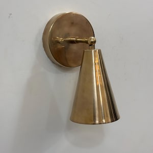 May include: A brass wall sconce featuring a circular backplate and a conical shade. The sconce has a pivoting arm. The finish is a warm, metallic gold. The sconce is mounted on a white wall.