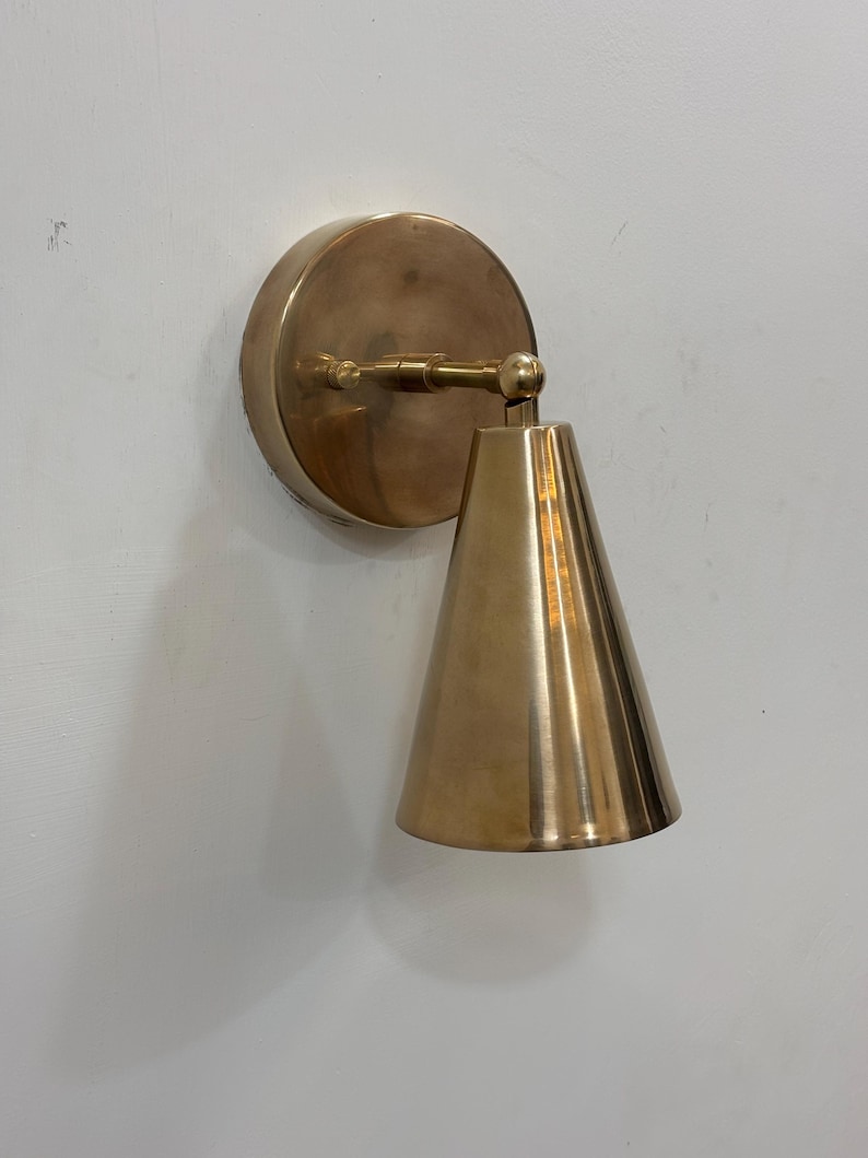 May include: A brass wall sconce featuring a circular backplate and a conical shade. The sconce has a pivoting arm. The finish is a warm, metallic gold. The sconce is mounted on a white wall.