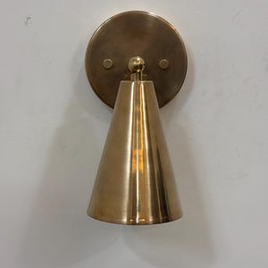May include: A brass wall sconce featuring a conical shade and a circular backplate. The sconce has a warm, metallic finish and is mounted on a white wall. The design is simple and modern, suitable for various interior styles.