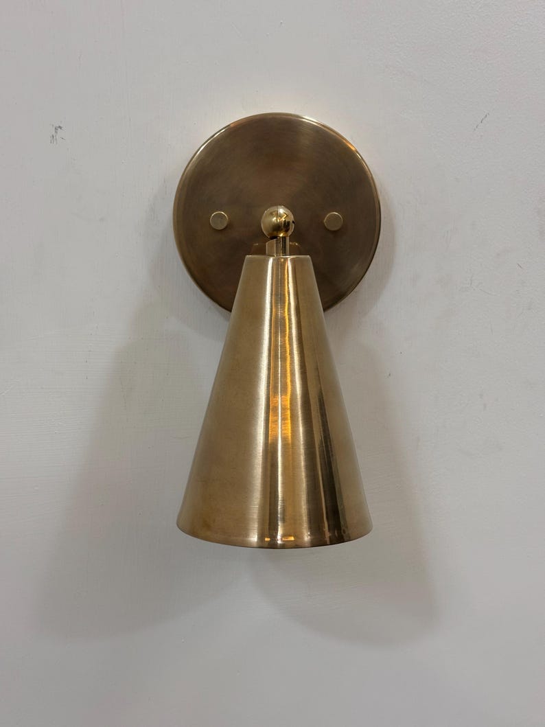 May include: A brass wall sconce featuring a conical shade and a circular backplate. The sconce has a warm, metallic finish and is mounted on a white wall. The design is simple and modern, suitable for various interior styles.