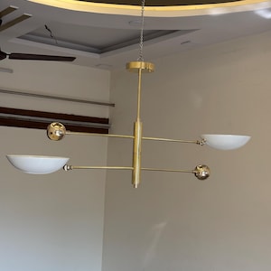 May include: A gold-toned modern chandelier with five light fixtures. The chandelier has a central rod with four arms extending out. Each arm has a round light fixture at the end. The light fixtures are white and have a bowl shape. The chandelier is hanging from a chain.