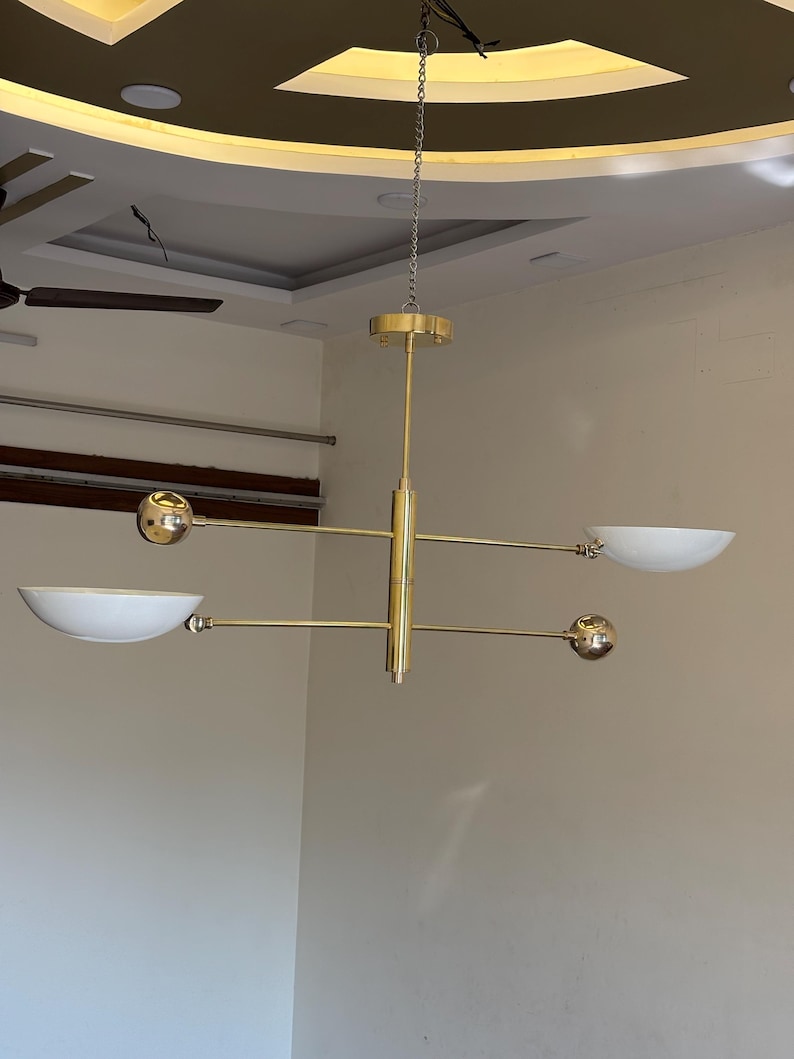 May include: A gold-toned modern chandelier with five light fixtures. The chandelier has a central rod with four arms extending out. Each arm has a round light fixture at the end. The light fixtures are white and have a bowl shape. The chandelier is hanging from a chain.