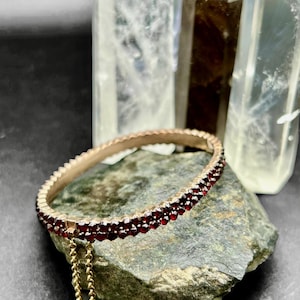 Puede incluir: Una pulsera rígida dorada con una doble fila de pequeñas piedras preciosas de color rojo intenso. La pulsera descansa sobre una piedra rugosa de color verde-gris, con puntas de cristal de cuarzo transparente en el fondo. Una delicada cadena dorada cuelga de la pulsera.