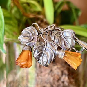 Op de afbeelding: Zilverkleurige broche met drie gestileerde bladeren en twee oranje-gele bloemhangers. De bladeren hebben gedetailleerde nerven en de bloemhangers hebben een doorschijnend uiterlijk. De broche is geplaatst tegen een vage groen-bruine achtergrond.