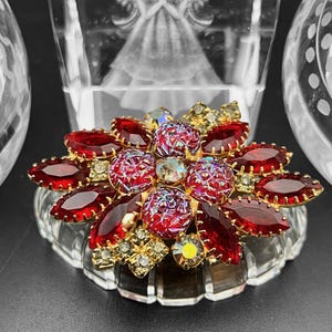 Op de afbeelding: Een rode en gouden broche met een bloemmotief. De broche is gemaakt van rode en heldere glazen stenen die in goudkleurig metaal zijn gezet.