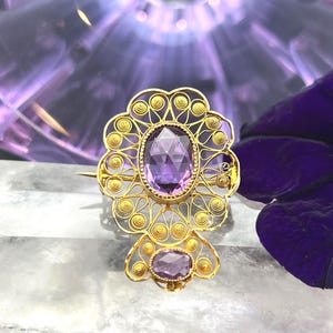 Può includere: Una spilla in oro caratterizzata da un intricato lavoro in filigrana con motivo a festoni. Il gioiello è incastonato con due pietre preziose ovali sfaccettate di colore viola, una più grande al centro e una più piccola nella parte inferiore.