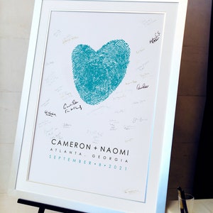 Blue thumbprint heart made from couple's fingerprints is framed and displayed on easel with wedding couple's names and wedding date and location. Wedding guest signatures surround heart on artwork.