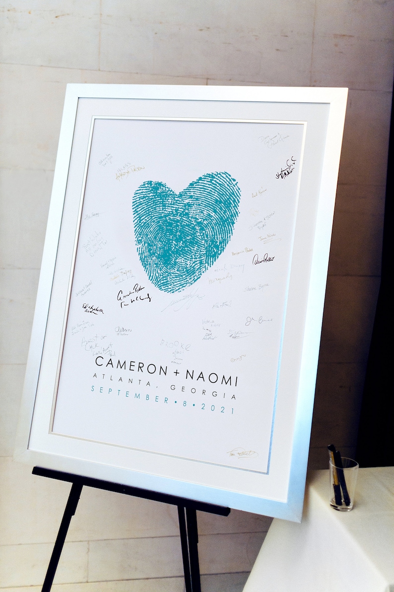 Blue thumbprint heart made from couple's fingerprints is framed and displayed on easel with wedding couple's names and wedding date and location. Wedding guest signatures surround heart on artwork.