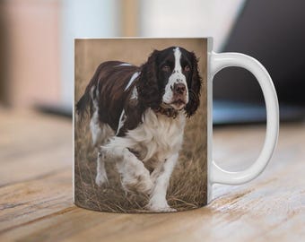 Könnte beinhalten: Eine weiße Keramiktasse mit einem braunen und weißen Springer Spaniel, der durch hohes Gras rennt. Der Hund schaut direkt in die Kamera.