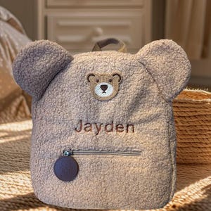 Peut inclure: Un sac à dos en peluche marron avec des oreilles d'ours, un visage d'ours brodé sur le devant et le nom "Jayden" brodé en dessous. Le sac à dos a une poche zippée et une étiquette en cuir marron avec un logo d'ours.