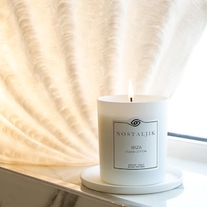 May include: White frosted glass candle with a black label that reads "Nostaljik Ibiza Clean Cotton Scented Candle Bougie Parfumée". The candle is lit and sitting on a white ceramic plate.