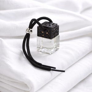 May include: A small, clear glass bottle with a black, patterned top and a black cord. The bottle is square-shaped and sits on a white surface. The cord has a silver bead and two dangling ends.