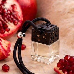 May include: A clear, square glass bottle with a black, decorative top and a black cord. The bottle is filled with a light-colored liquid. Red pomegranate fruit and seeds are scattered around the bottle.
