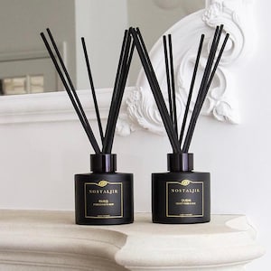 Könnte beinhalten: Zwei schwarze Glas-Diffusorflaschen mit schwarzen Stäbchen. Jede Flasche hat ein goldenes Etikett mit dem Wort "NOSTALJIK" und einem Städtenamen. Die Flaschen stehen auf einer weißen Oberfläche, mit einem weißen dekorativen Spiegel im Hintergrund.