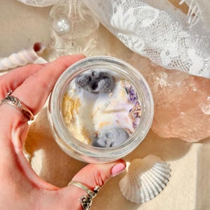 Puede incluir: Una pequeña vela en tarro de cristal transparente con base de cera blanca, adornada con flores secas, copos dorados y elementos decorativos grises. La vela se encuentra sobre un fondo de encaje, una concha y una figura de cristal transparente.