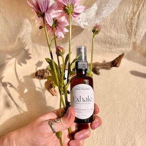 May include: A 2 fl oz amber glass spray bottle labeled Exhale botanical ritual mist sweet orange & sage by wild kalmia, held alongside a stem of pink flowers. The bottle features a black spray nozzle and a white oval label with black text.