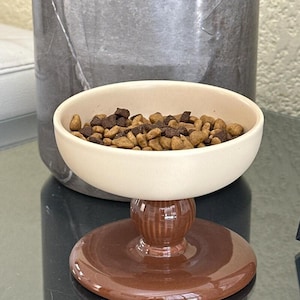 Puede incluir: Un cuenco de cerámica color crema para mascotas con un recipiente redondeado y liso, apoyado sobre una base de pedestal acanalada de color marrón oscuro brillante. El cuenco contiene una mezcla de croquetas secas marrón claro y oscuro.