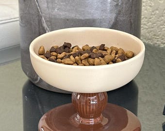 May include: A cream-coloured ceramic pet bowl with a smooth, rounded basin, supported by a glossy, dark brown, ribbed pedestal base. The bowl is filled with a mixture of light tan and dark brown dry pet food kibble pieces.