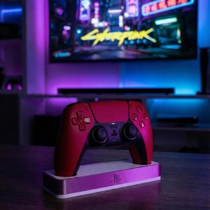 May include: A red and black wireless game controller resting on a rectangular white charging dock featuring a silver PlayStation logo. The controller has a central touchpad, two analog sticks, and directional buttons, set against a blurred gaming room.