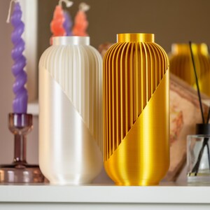 May include: Two decorative vases with a diagonal color split. One is ivory with a white diagonal, the other is gold with a gold diagonal. The vases have a ribbed design and are on a white surface.