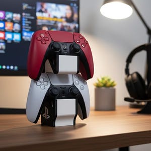 May include: A white and red game controller stand holding two game controllers. The top controller is red and black, and the bottom controller is white and black. The stand is on a wooden desk, with a computer monitor and headphones in the background.