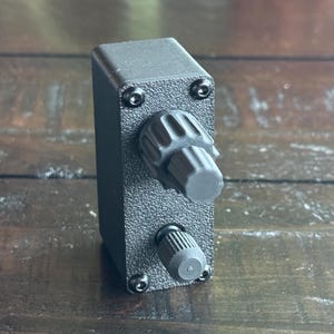 May include: A black box with two knobs, one large and one small. The box has a textured surface and is made of plastic.