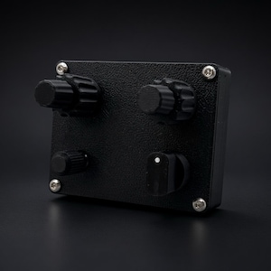 May include: A rectangular black electronic control box with a textured finish, featuring four black knobs on the front panel. Three knobs are cylindrical with ridged edges, while the fourth is a pointer knob with a small white dot indicator.