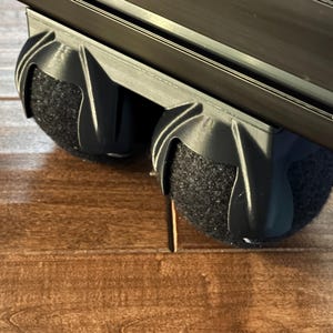 May include: Close-up of a black, 3D-printed component with two dark gray, spherical, felt-covered pads. The component is attached to a black, rectangular bar. The background is a wooden surface.