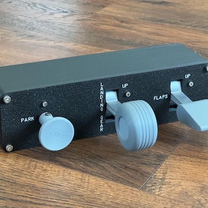 May include: A black control panel with gray levers and knobs. The panel has labels for "PARK", "LANDING GEAR", "UP", and "FLAPS". The levers and knobs are light gray. The panel is rectangular and has screws on the corners.