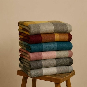 Op de afbeelding: Een stapel van zeven opgevouwen dekens in verschillende kleuren, waaronder geel, grijs, teal, roze en bordeaux. Elke deken heeft een gestreept patroon. De dekens zijn op een kleine houten kruk gestapeld.