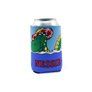 Può includere: Un portabevande blu con un'illustrazione a cartoni animati del mostro di Loch Ness, o Nessie, con un cappello scozzese. La parola "NESSIE" è stampata in rosso in basso. Lo sfondo è blu con onde bianche.