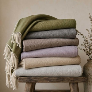 Puede incluir: Una pila de seis mantas dobladas con un patrón de espiga. Los colores incluyen verde oliva, gris carbón, marrón, morado claro, beige y gris claro. Cada manta tiene borlas de flecos de color crema a lo largo de los bordes laterales.