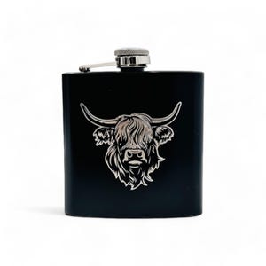 May include: Black stainless steel hip flask featuring a silver Highland cow illustration. The flask has a hinged cap and a silver top. The cow design includes curved horns and detailed fur.