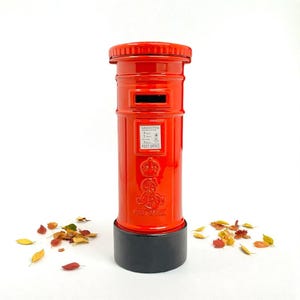 Può includere: Una cassetta postale cilindrica rosso vivo con base nera, che ricorda le classiche cassette postali britanniche. La cassetta postale presenta una fessura per la posta, una piccola targa informativa e uno stemma reale. Foglie autunnali sparse.