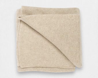 Könnte beinhalten: Eine gefaltete rechteckige Decke in einem einfarbigen hellen Beigeton. Das Textil weist eine feine, gleichmäßige Strickkonstruktion mit einer weichen, matten Textur auf der gesamten Oberfläche auf. Die Kanten sind mit einem schlichten Saum versehen.