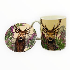 Peut inclure: Un mug en céramique et un dessous de verre assorti, ornés d'une illustration d'un cerf aux grands bois, entouré de fleurs violettes et de verdure. Le mug a un fond crème.